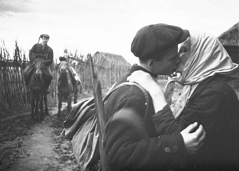„Kolchoznice M. E. Nikolajeva se loučí se syny, kteří se přidávají k partyzánům. Líbá mladšího Ivana, starší syn Semen je už na koni.“ Fotografie Michaila Trachmanna ze série pořízené v roce 1942 ve vesnici Kruglovo v Pskovské oblasti