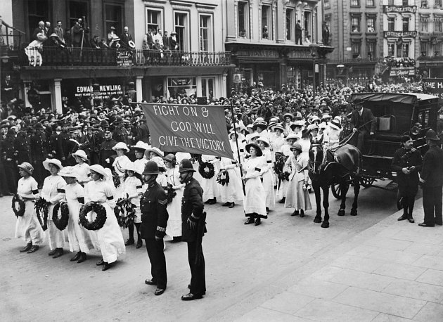 Část pohřebního průvodu během pohřbu Emily Davisonové 13. června 1913. Průvod sufražetek v bílém nese věnce a transparent s nápisem Bojujme dál a Bůh nám dá vítězství. Ulice lemovaly davy lidí
