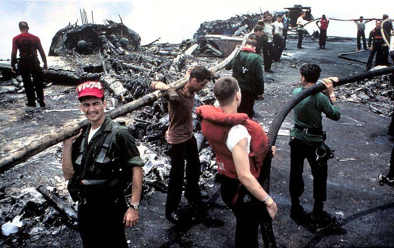 Zbylí vyčerpaní členové posádky Forrestalu stojí nad ohořelými troskami po palubním požáru, který si vyžádal životy 134 jejich druhů