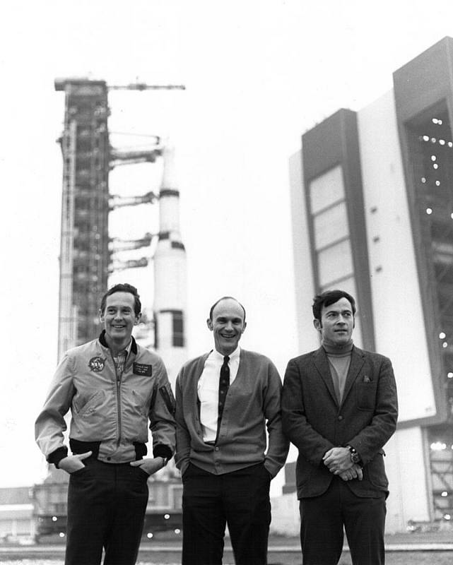 Posádka mise Apollo 16. Zleva Charles Duke, Thomas Mattingly a John W. Young. V pozadí za nimi raketa Saturn V, která je později vynesla do vesmíru.