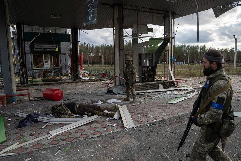 Ukrajinští vojáci procházejí kolem zničené čerpací stanice. Na zemi leží tělo jejich padlého spolubojovníka.