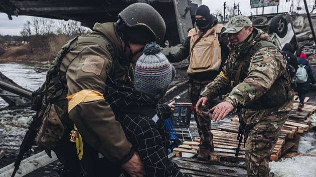 Ukrajinští vojáci ve městě Irpiň pomáhají civilistům překonat řeku. Most v pozadí Ukrajinci zničili, aby zpomalili postup ruských vojsk.