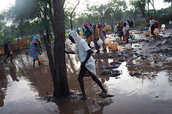 Ženy a děti se brodí vodou a bahnem, aby si naplnily své kanystry u distribučního místa pitné vody v táboře Batil. Silné deště a záplavy v táboře zhoršili už tak špatný stav vodních zdrojů a sanitace.