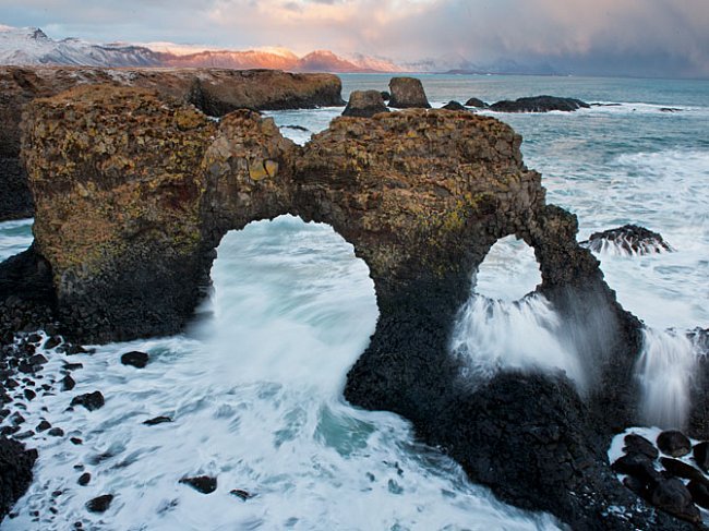 Zimní příboj se probíjí skalní branou, kterou vyhloubil v čedičových útesech v Arnarstapi na poloostrově Snæfellsnes. 