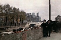 Dva muži stojí u drobností prodávaných na nábřeží Seiny ve dvacátých letech minulého století, s katedrálou Notre Dame v pozadí.