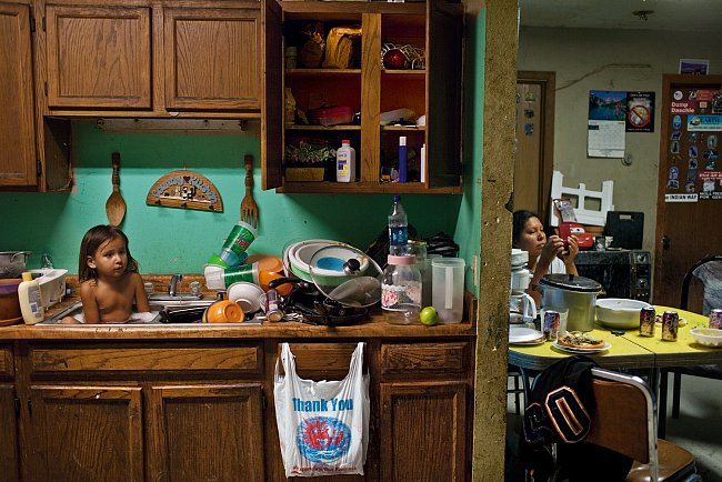 Tříletý C. J. Shot se koupe mezi nádobím. V souladu s představou Oglalů o tiospaye – jednotě a spolupráci členů rozšířené rodiny – jsou jejich domovy často přelidněné. K tomuto stavu přispívá i kritic