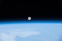 Měsíc, taneční partner Země, vyfotografovaný z Mezinárodní vesmírné stanice vykukuje nad rozostřenou modrou vrstvou atmosféry.