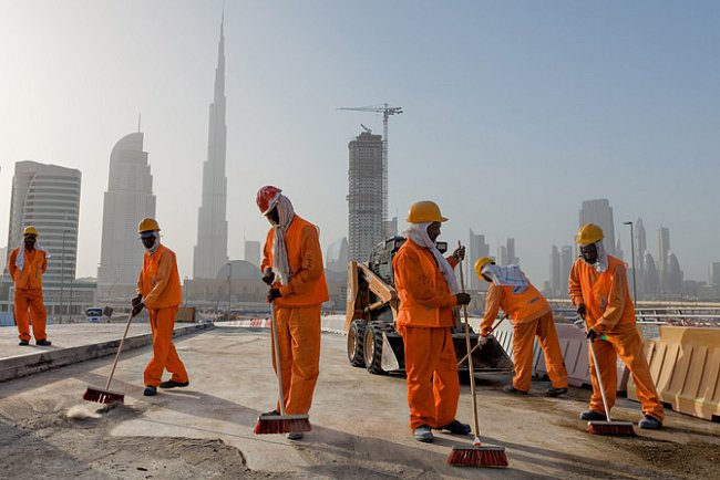 Nejvyšší budova na světě Burdž Chalífa se tyčí jako vzdálený bajonet nad dělníky zametajícími staveniště. Pocházejí většinou z Pákistánu a Indie.