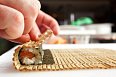 4. Nyní sushi zarolujte. Podložku chyťte zespoda a sushi začněte pomalu pevně zavinovat. Důležité je na sushi přehnaně netlačit, ale jen lehce zamotat. Rolku přimáčkněte z boku a seshora, aby dostala tvar a konec řasy se dobře přilepil. 