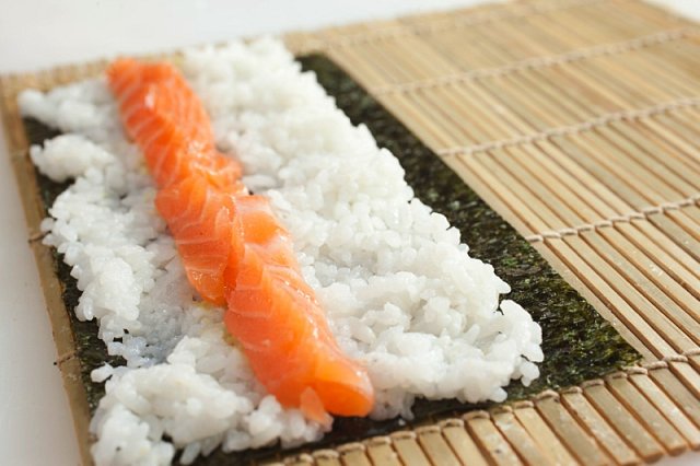 2. Vezměte 75 g uvařené sushi rýže a naneste ji na řasu. Na vzdálenějším okraji řasy nechte volný centimetrový proužek – tam se pak řasa po zarolování slepí k sobě (řasu nenamáčejte). Rýži lehce potřete wasabi, posypte sezamovým semínkem a poklaďte v jedn