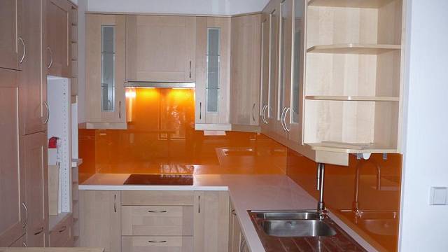 Skleněný obklad nemusí být výsadou jen moderních nebo minimalistických kuchyní! V teplé zářivé oranžové barvě je tmavému a elegantnímu ořechovému dřevu stylové kuchyně  rovnocenným partnerem. Různé typy a barvy skel vhodných pro skleněné obklady dodává...