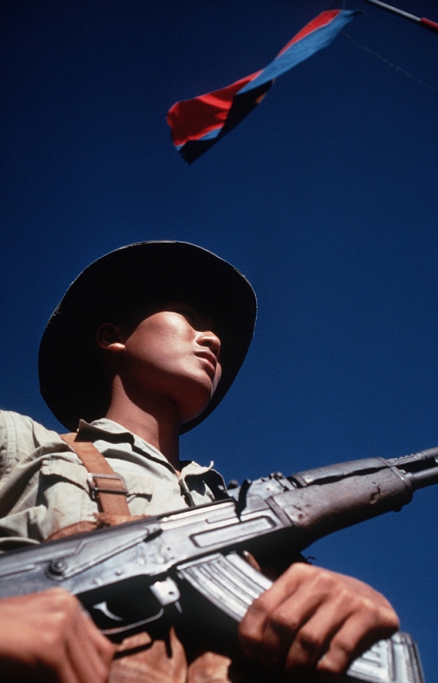 Voják Viet Kongu stojí pod vlajkou Viet Kongu a nese svou pušku AK-47.