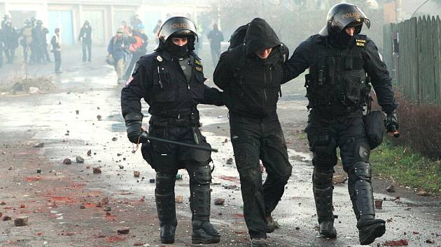 Policisté odvádějí jednoho z demonstrantů při listopadových nepokojích v Janově. Na zemi leží kusy cihel a kamení z rozebraných plotů při cestě.