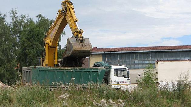 Bagr složila z podvalníku stavební firma na pozemku někdejší továrny o víkendu. V pondělí se lžíce stroje zakousla do země a mění zaplevelenou parcelu k nepoznání.