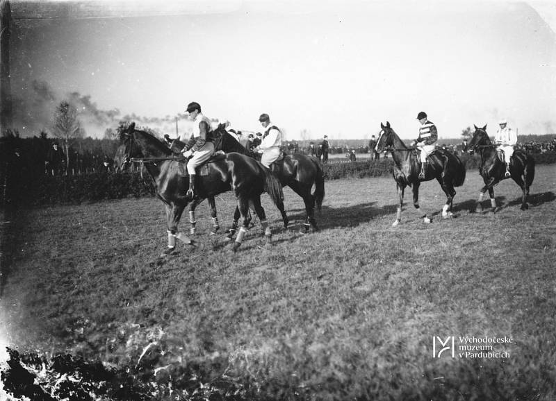 Velká pardubická, 4 dostihoví závodníci míří na start, v pozadí komíny Pardubic.