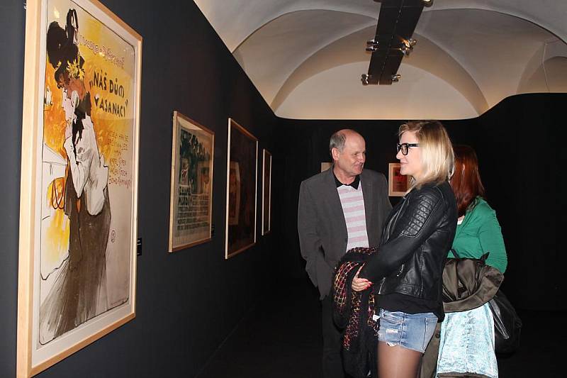 Fotografie zachycuje návštěvníky výstavy Karikatura a její příbuzní před plakátem Luďka Marolda ke hře Náš dům v asanaci
