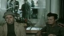 Muž z Londýna. Na snímku je vyslýchán herec Václav Babka (Fifka), za ním průhledem v okně pracují celníci