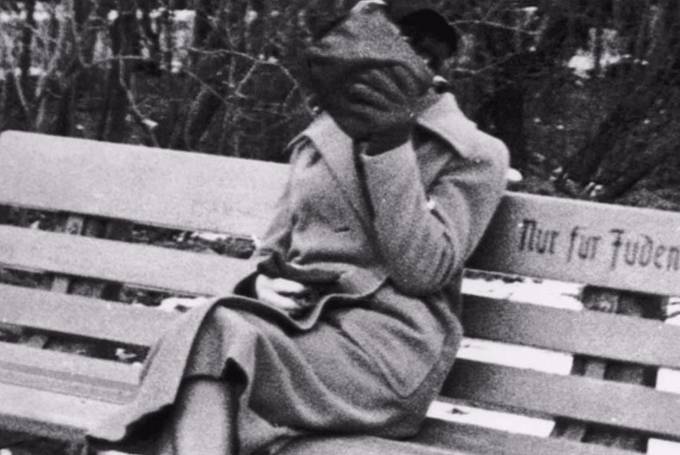 Židovská žena, která si halí tvář, aby nebyla poznat. Na lavičce je nápis "Pouze pro Židy". V roce 1938, z kterého je fotka, byli Židé striktně separováni od nežidovského obyvatelstva.