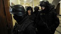 Ozbrojená eskorta chránila jednu z obžalovaných žen, která získala status spolupracující obviněné. Na sobě měla neprůstřelnou vestu a tvář jí zakrývala maska. 