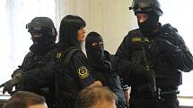 Ozbrojená eskorta chránila jednu z obžalovaných žen, která získala status spolupracující obviněné. Na sobě měla neprůstřelnou vestu a tvář jí zakrývala maska. 